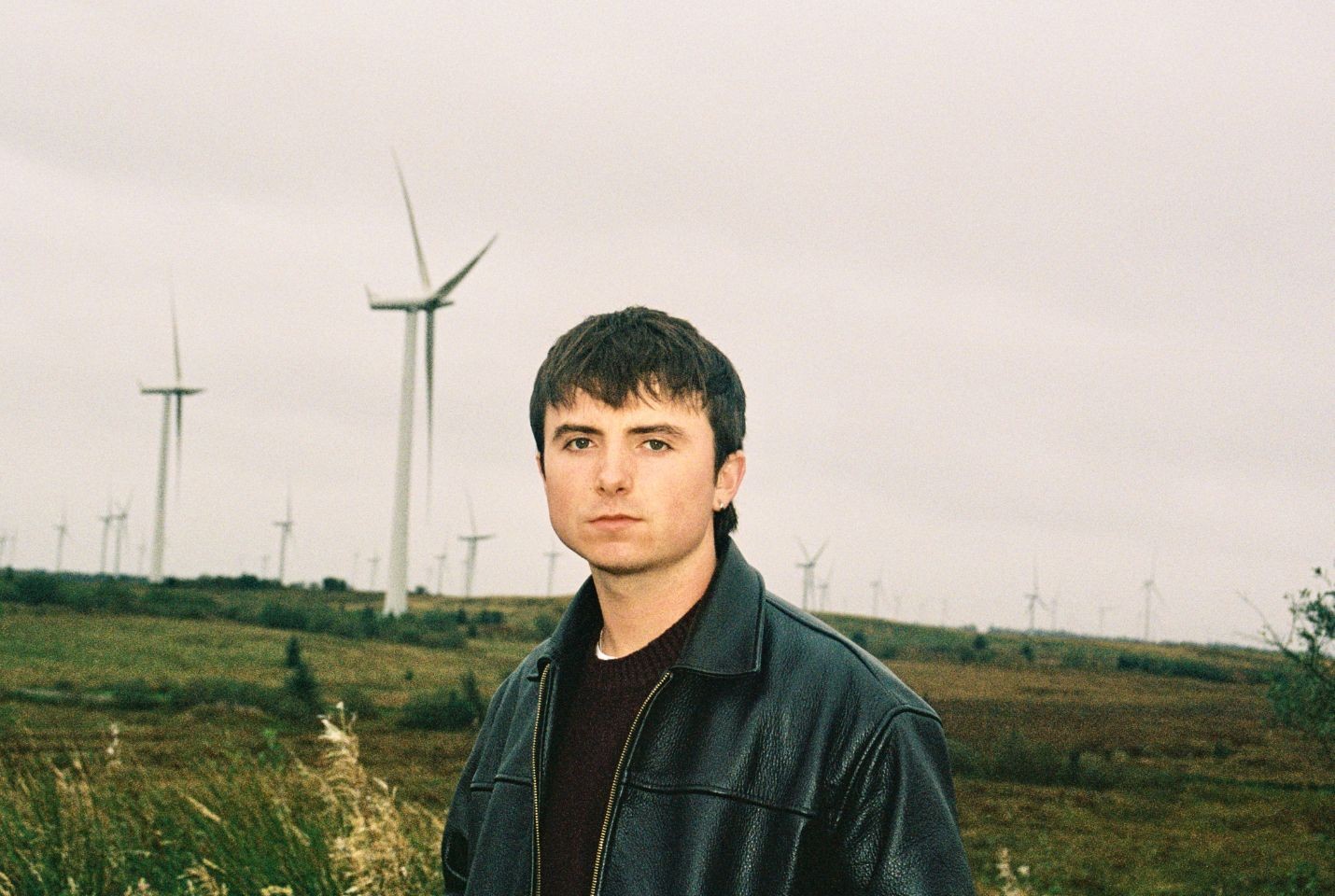 A guy in a brown jumper and black leather jacket stood in a field in front of wind turbines